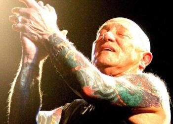 Gary "Angry" Anderson of Rose Tattoo on stage, eyes closed, clapping his tattooed arms overhead under a spotlight.