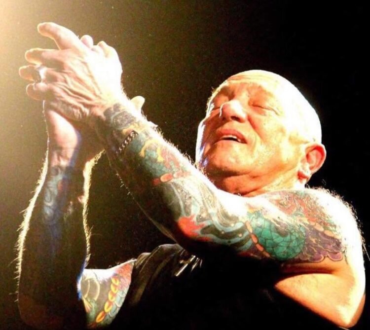 Gary "Angry" Anderson of Rose Tattoo on stage, eyes closed, clapping his tattooed arms overhead under a spotlight.