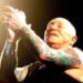 Gary "Angry" Anderson of Rose Tattoo on stage, eyes closed, clapping his tattooed arms overhead under a spotlight.