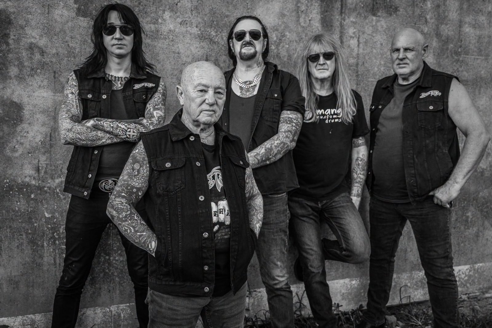 Black-and-white promo photo of Rose Tattoo, five tattooed band members in denim vests posing against a weathered concrete wall.