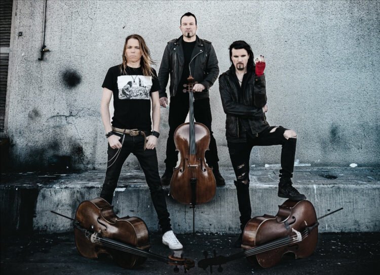 Apocalyptica posing in black clothing against a grey concrete wall, with three cellos placed around them