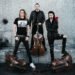 Apocalyptica posing in black clothing against a grey concrete wall, with three cellos placed around them