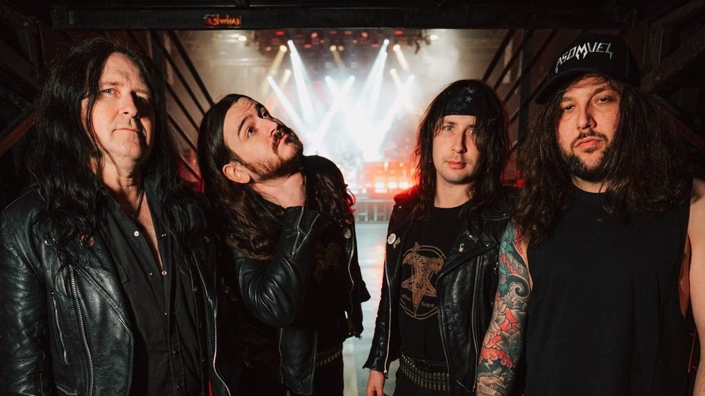 Asomvel in leather jackets posing backstage in front of a brightly lit stage.