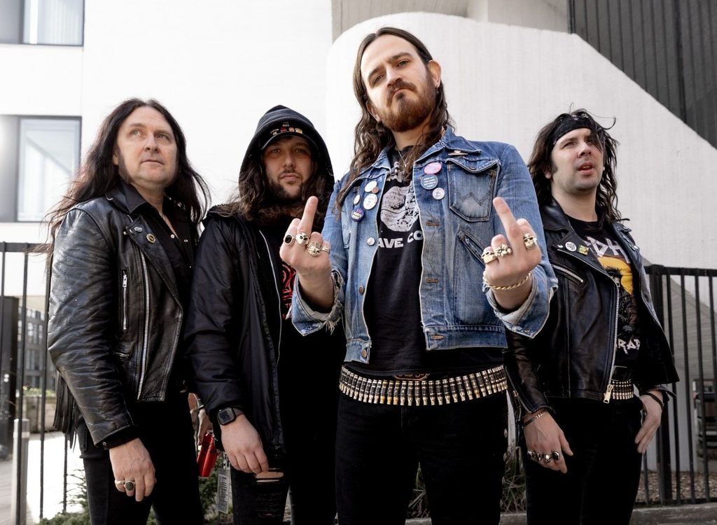Asomvel posing outdoors in front of a white building, with one member in a denim jacket raising both middle fingers.
