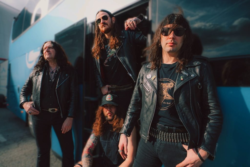 Asomvel in leather jackets and sunglasses posing beside a blue tour bus.