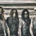 Belphegor band members in corpse paint and leather vests posing in front of a corrugated metal wall