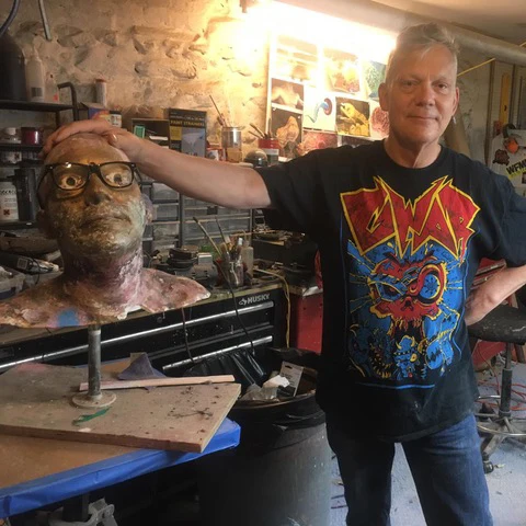 Chuck Varga in a GWAR T-shirt resting his hand on a sculpted head wearing glasses in his art studio.