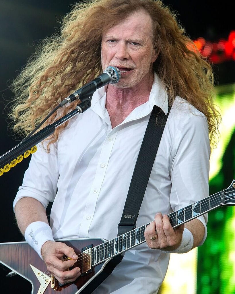 Dave Mustaine of Megadeth singing into a microphone while playing a V-shaped guitar on stage, wearing a white shirt