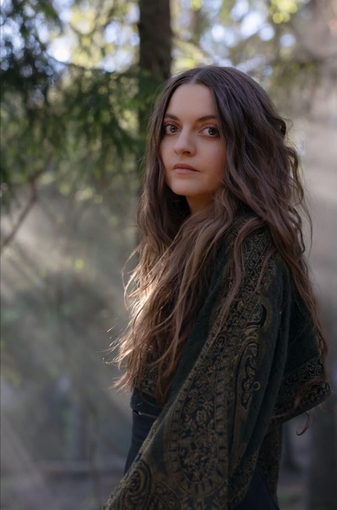 Katrine Stenbekk of Kalandra with long brown hair and a patterned shawl, standing in a sunlit forest