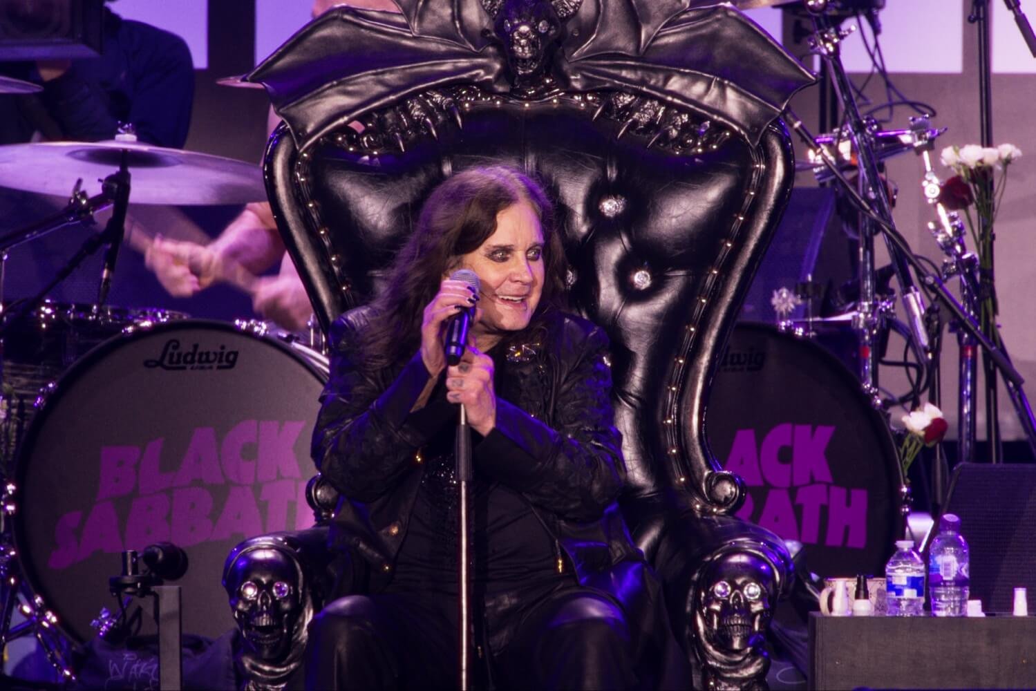 Ozzy Osbourne singing from a black skull-armed throne in front of Black Sabbath drum kits.