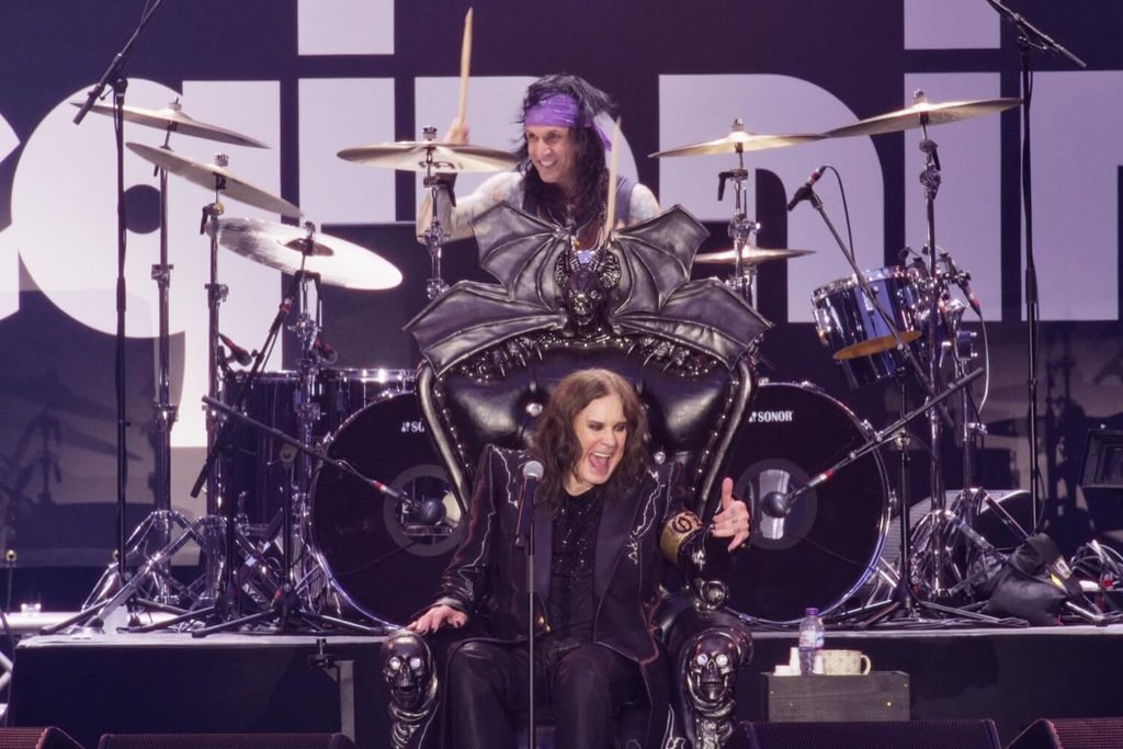 Ozzy Osbourne laughing on a black bat-winged throne on stage, with a drummer playing behind him