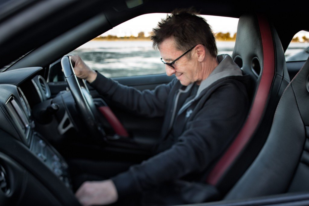 Phil Rudd smiling behind the wheel of a ferrari sports car, wearing glasses and a dark hoodie.