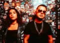 Electron in front of a sticker-covered wall, her in a skeleton-print jacket and him in sunglasses and chains