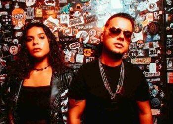 Electron in front of a sticker-covered wall, her in a skeleton-print jacket and him in sunglasses and chains