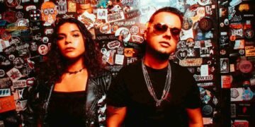 Electron in front of a sticker-covered wall, her in a skeleton-print jacket and him in sunglasses and chains