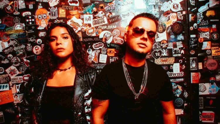 Electron in front of a sticker-covered wall, her in a skeleton-print jacket and him in sunglasses and chains