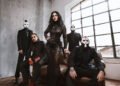 Lacuna Coil posing in black outfits and white face paint in a loft with an arched window, the singer standing at the center in a black lace gown.