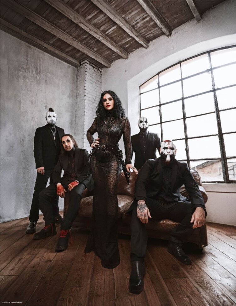 Lacuna Coil posing in black outfits and white face paint in a loft with an arched window, the singer standing at the center in a black lace gown.