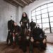 Lacuna Coil posing in black outfits and white face paint in a loft with an arched window, the singer standing at the center in a black lace gown.