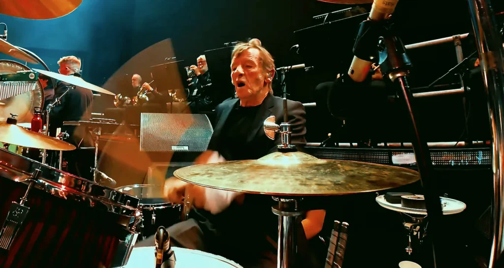 Phil Rudd playing drums on stage, with an orchestra's brass section behind him