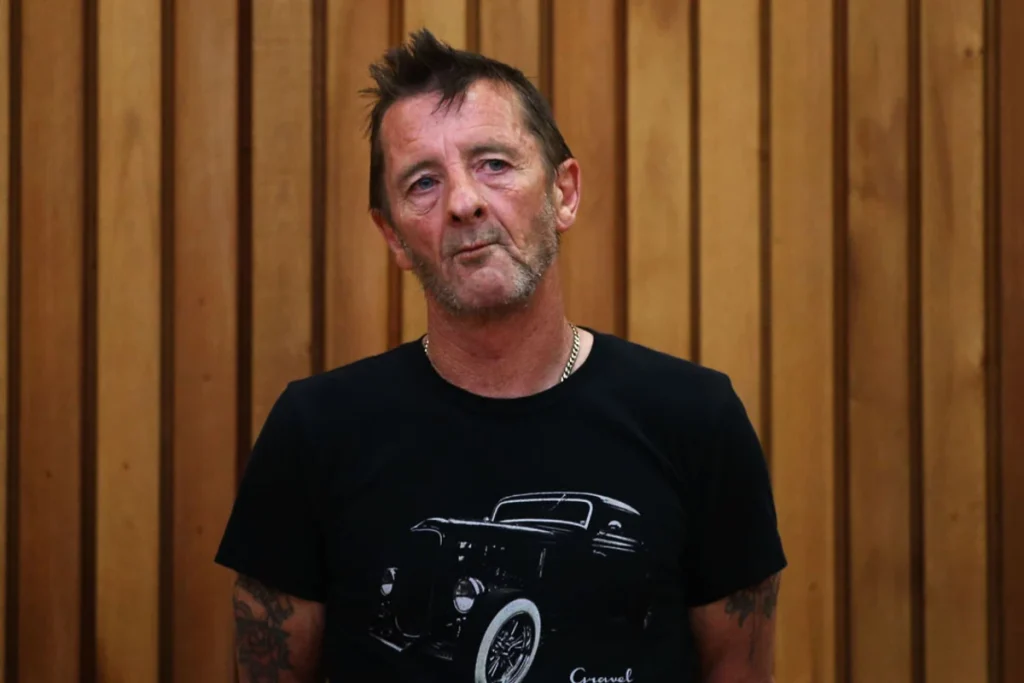Phil Rudd in a black hot rod T-shirt standing in front of a wooden panel wall